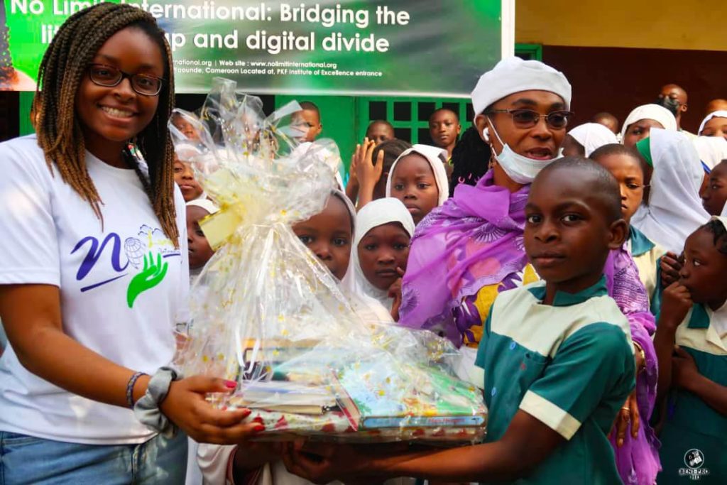 No Limit International Co-Founder Orlyanka Tantchou holds a bag of book donations alongside a young school student. Behind them are a group of more students and a school leader.