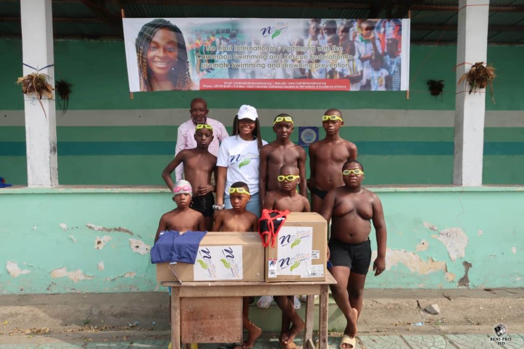 No Limit International Co-Founder Orlyanka Tantchou stands with a group of young swimmers and a swimming education leader. In front of them on a table are boxes of swimming equipment donations, provided by No Limit International and A3 Performance.