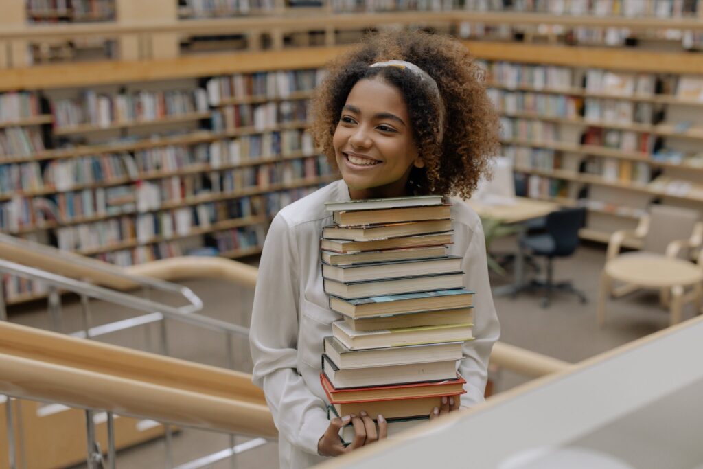 A girl with kinky, curly hair carrying a pile of books in the library.