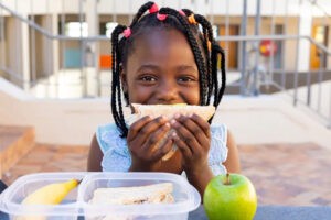A girl sits with her meal outside. She is holding half a sandwich up to her mouth. In front of her is another sandwich half, a banana, and a green apple.