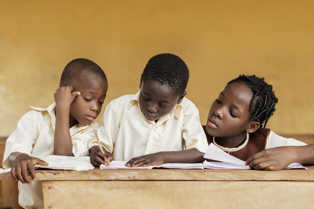 Three school children gather at a desk with notebooks. The two children on the left and right look at the notebook in the middle, which is being written in by the child in the middle.