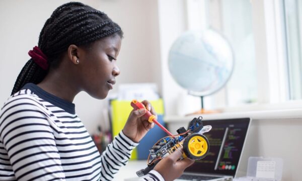 A black female teenager sits at her desk while working on a STEM project with a screwdriver in hand. On the desk is an open laptop with code on the screen as well as other office supplies.