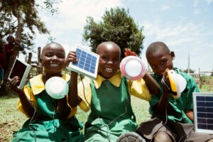 A group of three schoolchildren in Kenya sit outside and hold up solar technology devices, including solar panels and other clean energy equipment.