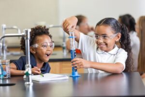 Two children perform a science experiment in a classroom laboratory. Both children are wearing safety glasses and smiling as they sit at the lab table.