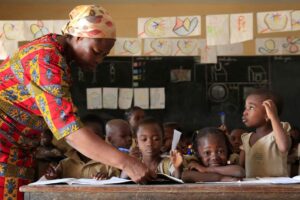 A teacher in an African classroom stands over a group of students sitting at a desk. She holds a notebook belonging to one of the students.